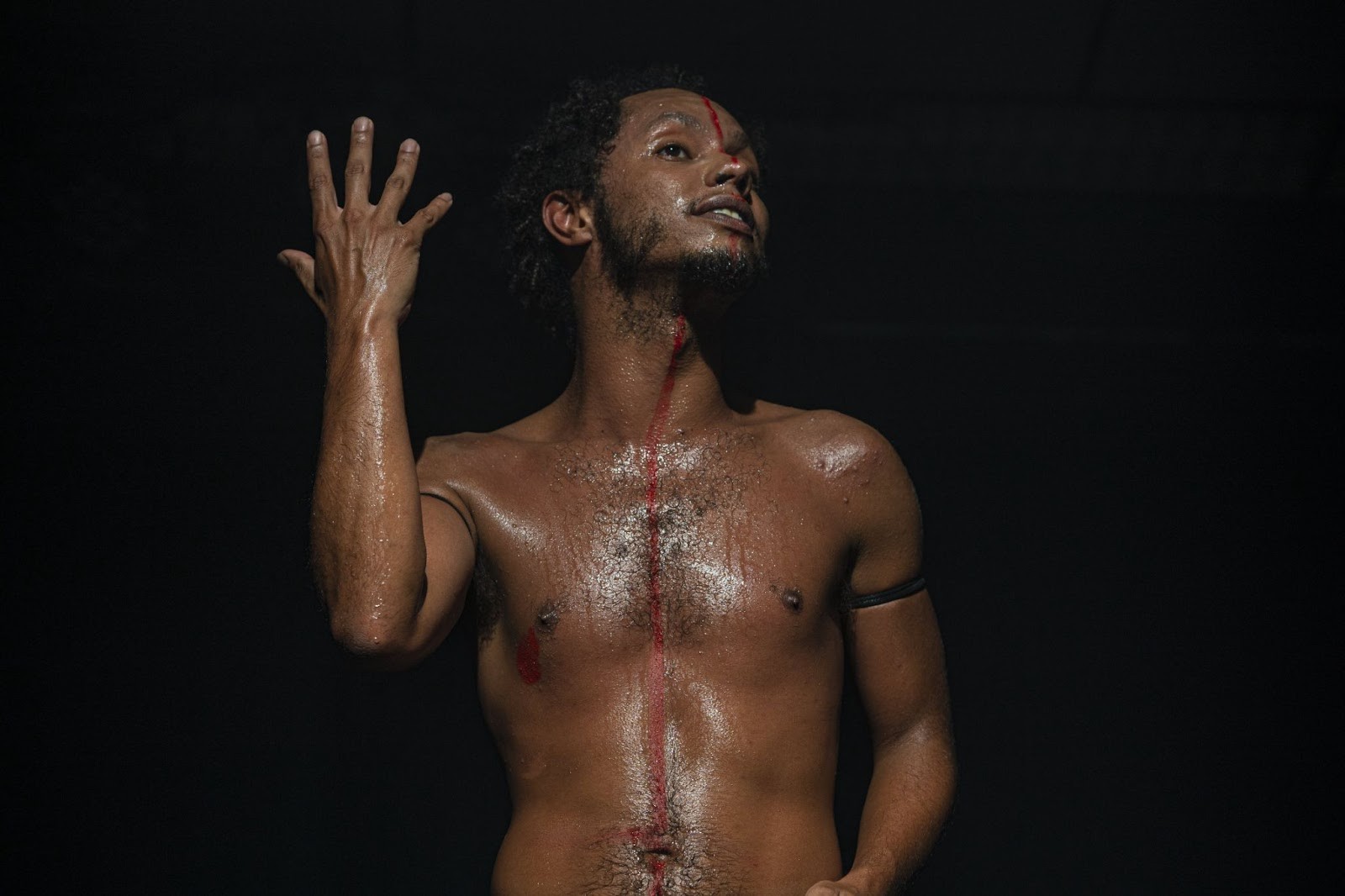 Homem negro, sem camisa, com o corpo suado e uma linha vermelha pintada do rosto ao umbigo. Ele ergue a mão esquerda, com os dedos abertos, e olha para cima, com expressão de súplica ou êxtase, em um fundo escuro