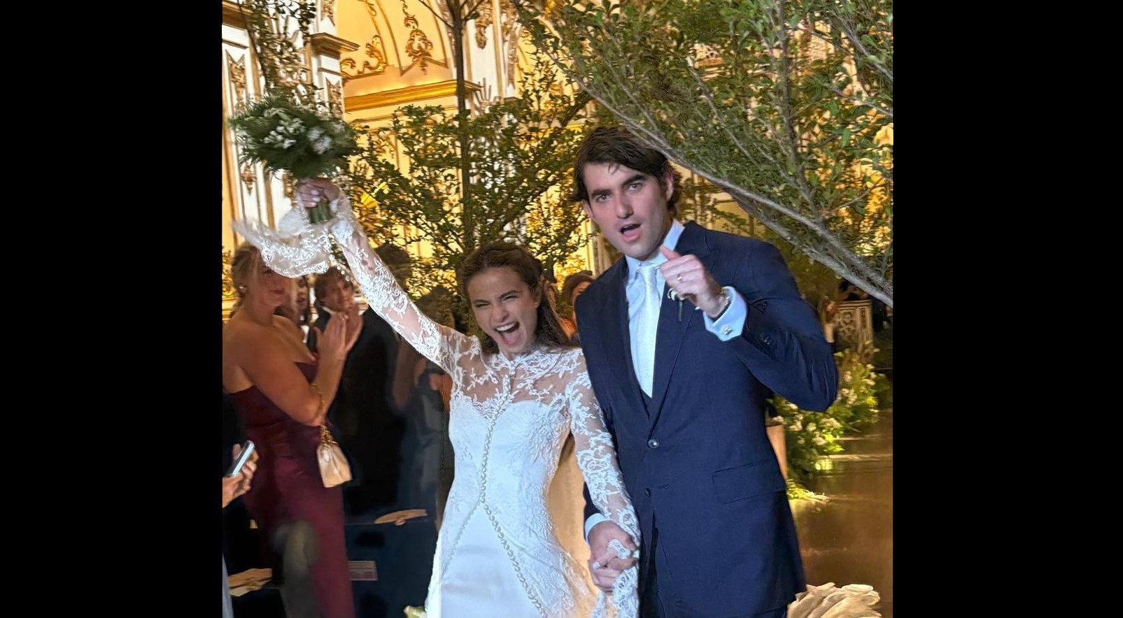 Noiva sorridente em vestido de renda branco com buquê erguido, de mãos dadas com noivo de terno azul-marinho, ambos comemorando em festa de casamento com decoração verde e dourada