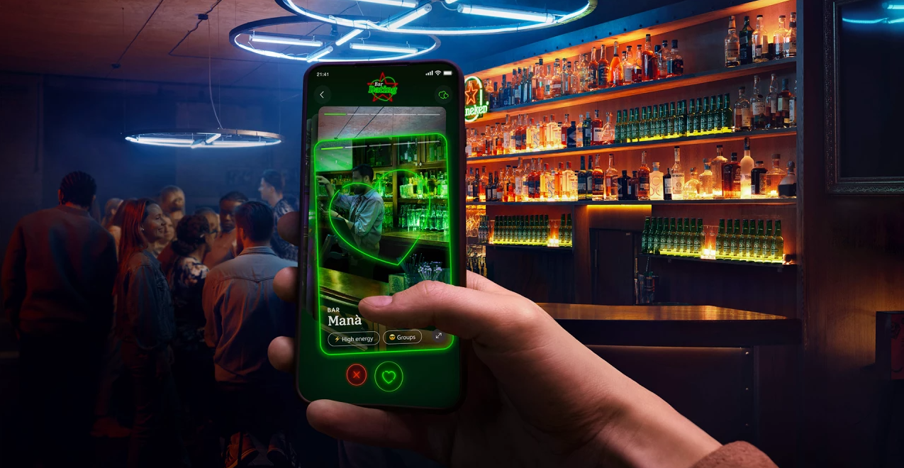 Mão segura smartphone com aplicativo de realidade aumentada exibindo o interior de um bar chamado Manà, com um bartender e garrafas iluminadas em verde. Ao fundo, um bar real com pessoas, balcão e prateleiras cheias de garrafas coloridas, sob luzes neon azuis