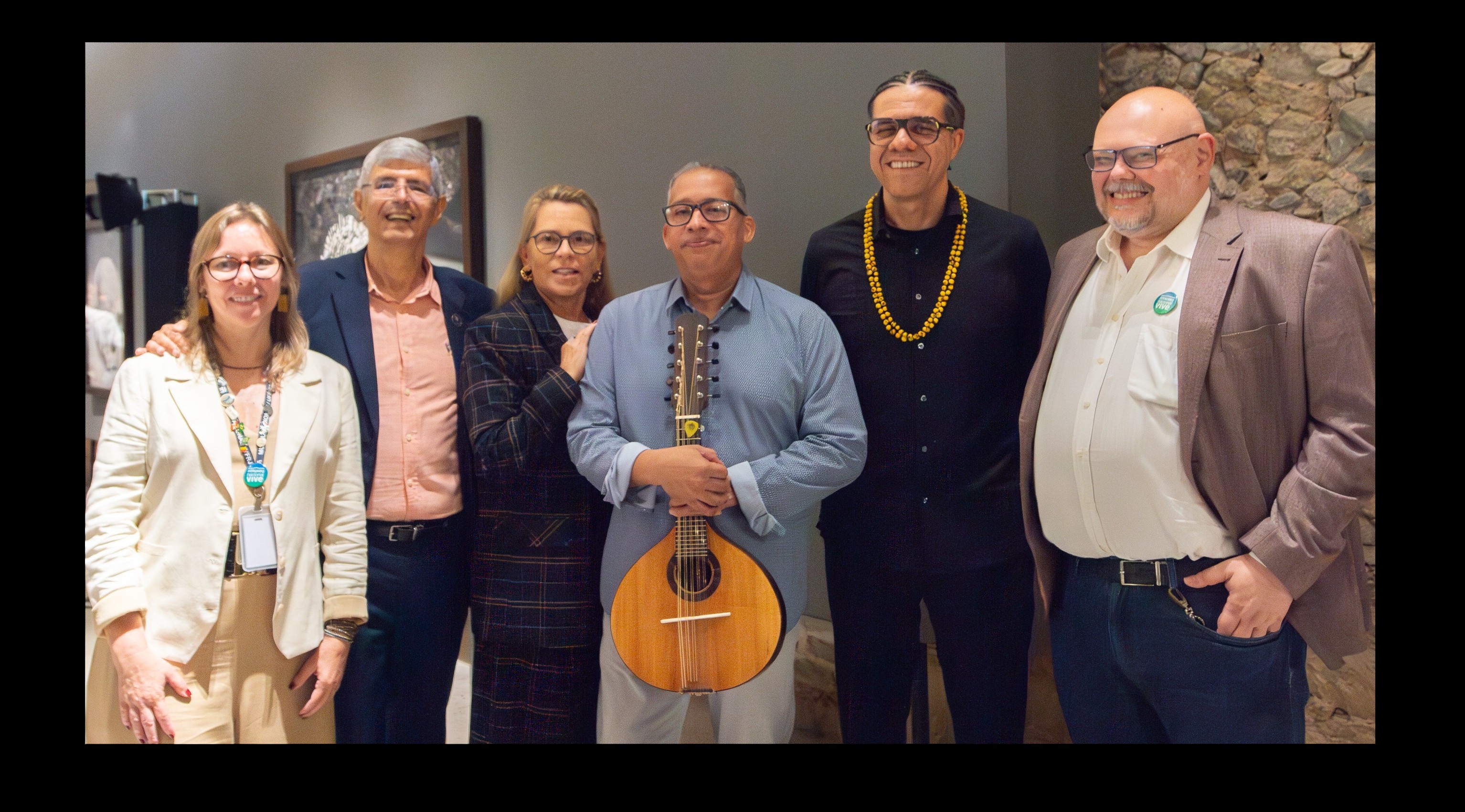 Seis pessoas sorridentes posam para foto. Uma mulher de blazer creme e óculos, um homem de blazer azul e camisa salmão, uma mulher de blazer xadrez, um homem de camisa azul segurando um bandolim, um homem de camisa preta e colar amarelo, e um homem de blazer marrom e camisa branca com broche verde