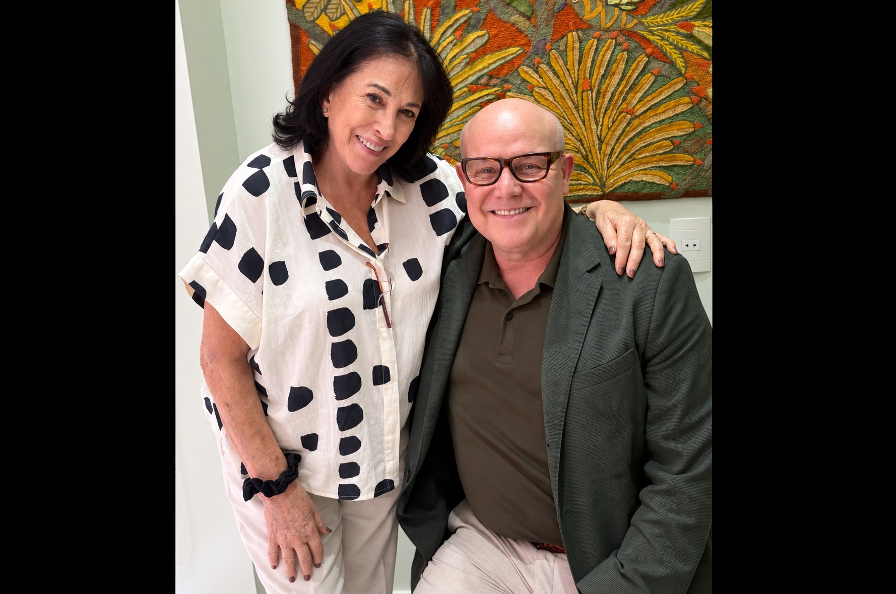Mulher de cabelos escuros e blusa branca com manchas pretas sorri ao lado de homem careca de óculos, blazer verde-escuro e camisa polo marrom. Ele tem o braço dela sobre o ombro. Ao fundo, uma tapeçaria colorida com motivos de folhas